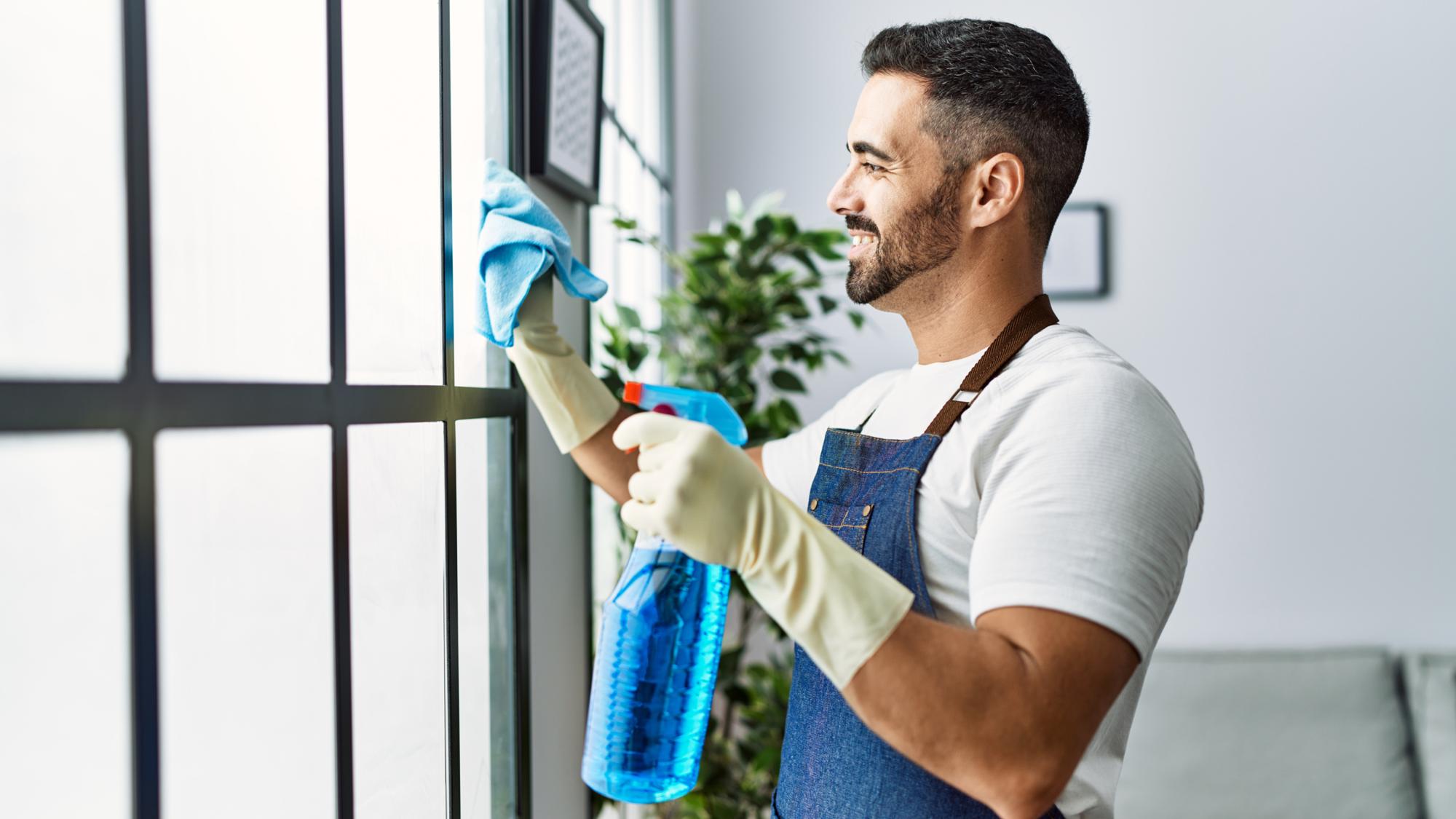 A man wearing yellow cleaning gloves, a white shirt, and a blue apron is cleaning a window with a spray bottle and a cloth. He is smiling and appears focused on the task. Indoor plants and a soft couch are visible in the background.